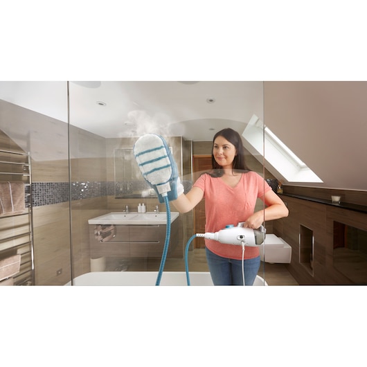 A person using a BLACK+DECKER steam cleaning device with a mitt attachment to clean a glass shower door in a modern bathroom.