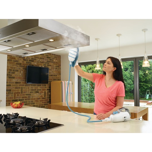 A person uses a BLACK+DECKER steam cleaning device (model BHSM1615DAM) to clean a kitchen exhaust hood. The device emits steam to remove dirt and grease. The kitchen has a modern design with brick walls, large windows, and a gas stove.