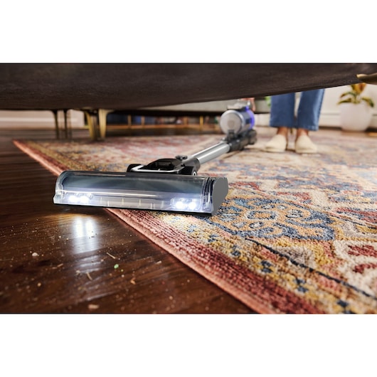 BLACKDECKER BSV520BRP cordless vacuum cleaning under sofa on hardwood floor and patterned rug, LED lights on head.