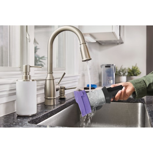 Photo of a BLACKDECKER BSV520BRP vacuum filter with purple cap being rinsed under a kitchen faucet in a stainless steel sink.