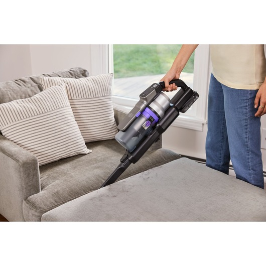 BLACKDECKER BSV525BRP cordless stick vacuum cleaning a grey upholstered chair in a living room.
