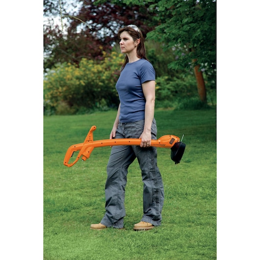 A person standing outdoors on a grass lawn, holding an orange BLACK+DECKER GL360SB grass trimmer. The person's face is blurred and they are wearing safety glasses, a blue t-shirt, grey pants, and brown shoes.