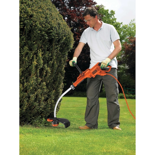 A person using a BLACK+DECKER electric grass trimmer to trim the lawn and edges near a hedge, outdoors in a garden.