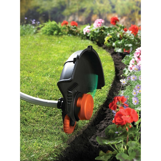 A BLACK+DECKER garden trimmer being used to edge a lawn next to a colorful flowerbed with red and purple flowers.