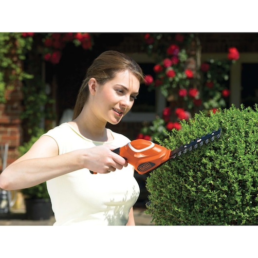 Photo of BLACKDECKER GSL700 cordless shrub trimmer in use, orange body and black blades trimming green bush.