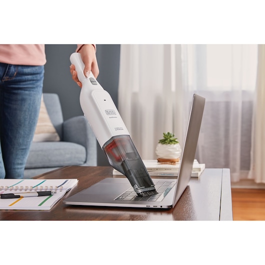 BLACK+DECKER HLVC315B11 handheld vacuum being used to clean a laptop keyboard on a desk in a home setting.