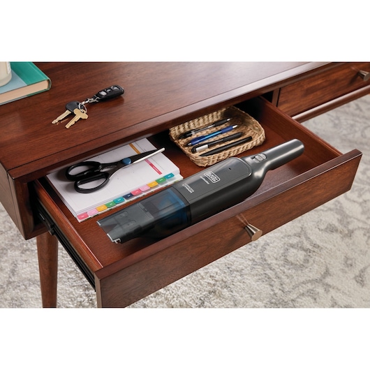 Photo of BLACK+DECKER HLVC320J11 handheld vacuum in an open wooden desk drawer with office supplies.