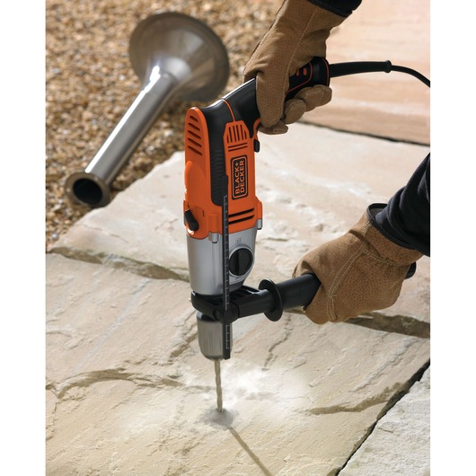 A person wearing protective gloves is using a BLACK+DECKER electric drill to drill into a stone surface, with dust rising from the hole. Some construction equipment is visible in the background.