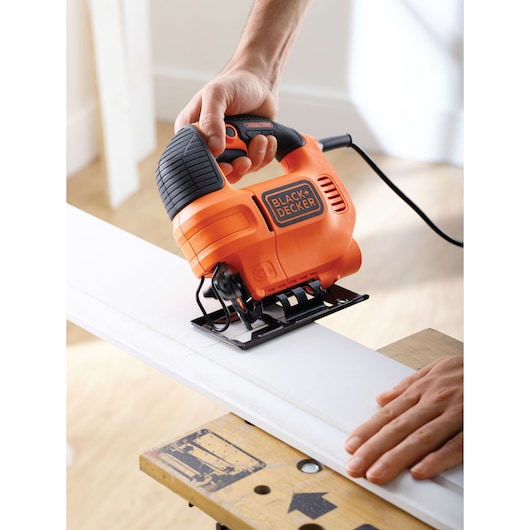 Photo of BLACK+DECKER KS701EK jigsaw cutting white wood, handheld, black and orange tool in use indoors.