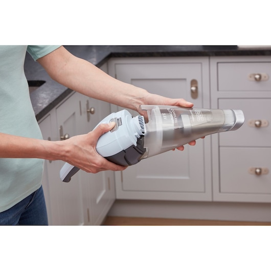 BLACK+DECKER NVC215W handheld vacuum cleaner with clear dust container being emptied in a kitchen setting.