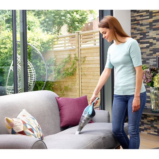 BLACKDECKER NVC220WBC handheld vacuum cleaner used to clean a grey sofa in a living room.