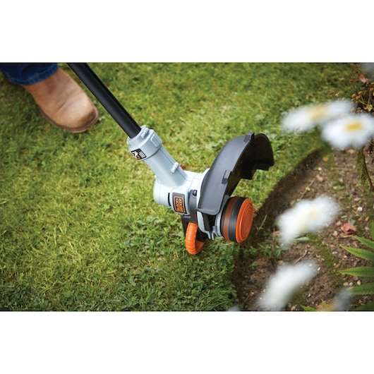 A close-up of a BLACK+DECKER grass trimmer being used to edge a lawn, showing the cutting head near a garden bed.