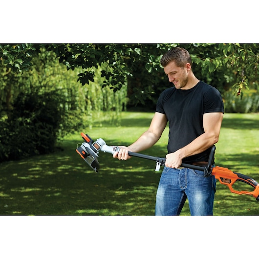 A person using a BLACK+DECKER STC1840PC grass trimmer in a garden. The person's face is blurred. The orange and black trimmer is held with both hands and the garden is green and sunny.